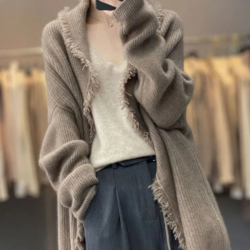 A person stands at the center, wearing a Lacatang Shop's Autumn Winter New Fashion V-neck Long Sleeve Solid Tassels Cardigan Sweater in beige, draped over a light top and tailored gray pants. The loose, oversized cardigan features rolled-up sleeves. Blurred clothing on hangers creates background images that gently harmonize with the outfit's neutral tones.