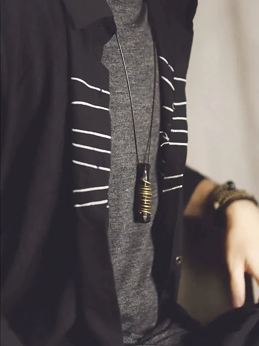 A person wearing a dark shirt with white stripes, layered over a gray t-shirt, showcases their style while highlighting the quality of their Original Design Ebony Wood Pendant from Lacatang Shop. This male temperament retro Chinese ethnic style necklace, featuring a stunning black and gold design on a woolen chain, exudes confidence in both fashion and care as they rest a hand on their leg.