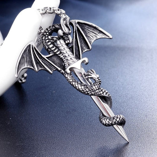 The Flying Dragon With Sword Necklace by Yellow Pandora features a pendant of an intricately designed flying dragon wrapping around a pointed blade, lying on a dark surface. The detailed alloy dragon's scales and wings are meticulously crafted, and the pendant hangs from a chain, supported from behind by a white object.