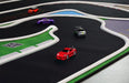 Four tiny cars race on Lacatang Shop's Turbo Drift Remote Control Car Track for Big and Small Scenes, featuring green grass-like borders. Red, orange, purple, and gray cars circle curved lanes, with a small blue pond-like section adding to the drifting excitement.