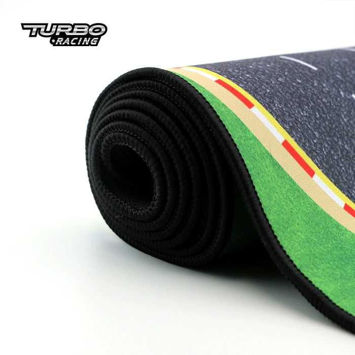 Close-up of a rolled-up racing-themed mouse pad. The design showcases a tarmac road with yellow and red markings, set against a green backdrop, resembling the Turbo Mini Drift Scene Remote Control Car Track by Lacatang Shop. The "Turbo Racing" logo appears in the top left corner.