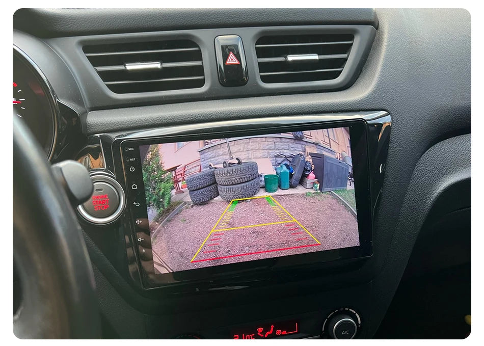 The Lacatang Shop's Develuck HD 1080P Car Rear View Camera displays a driveway with tires, a broom, and garbage bins. Parking guidelines are clear on screen, while the dashboard shows a hazard button above the display. The camera's waterproof feature maintains clarity in any weather.