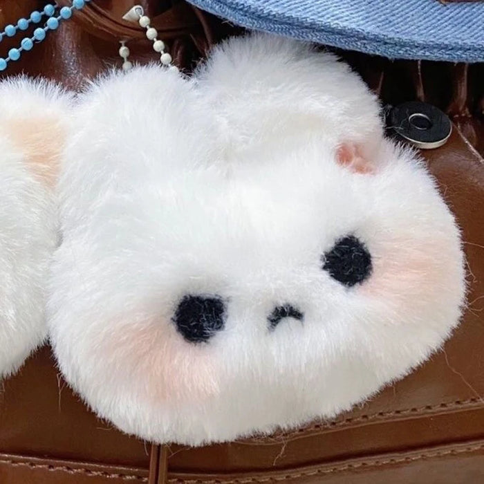 A fluffy white Cartoon Plush Rabbit Doll Toy Keychain from Lacatang Shop, crafted from high-quality plush material and shaped like a round-faced rabbit with small black eyes and a tiny frown, is attached to a brown leather bag. The plush has light pink ears and cheeks, giving it a cute, slightly sad expression.