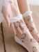 A hand adjusts a sheer sock, embroidered with pink roses and green leaves. The Lacatang Shop's Vintage Floral Lace Ruffle Socks, designed in a Kawaii Harajuku style, embellish the foot against wooden flooring, with a white garment sleeve partly visible.