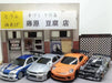 Four models from Lacatang Shop, which include a silver sports car, a white sedan with a spoiler, an orange Porsche, and a black race car, are parked in front of a miniature building that features a sign with Japanese text. Each 2.4G RC Drift Car 1/43 by Lacatang Shop promises high speed and four-wheel drive capabilities for thrilling races.