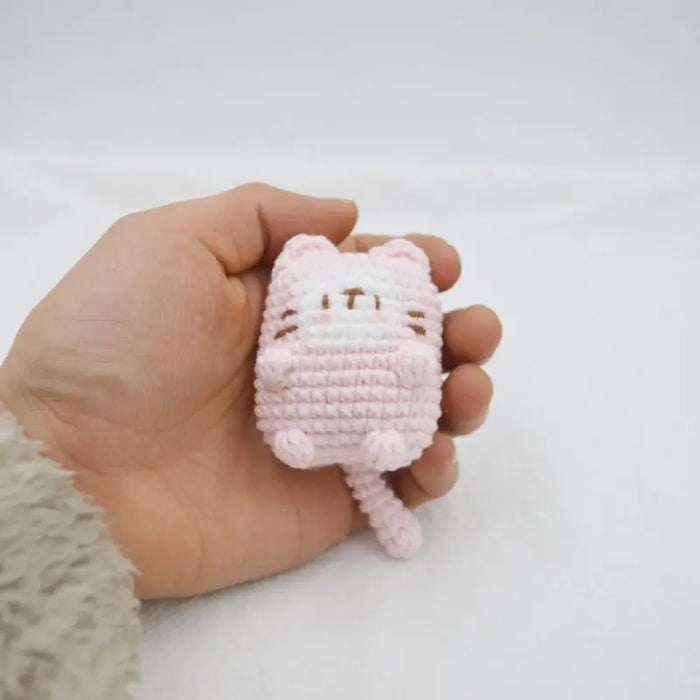 A hand is holding a small, crocheted, light pink cat from the Lacatang Shop's Cute Cat Crochet Keychains collection. The adorable cartoon cat doll has closed eyes and tiny limbs, fitting snugly in the palm and appearing soft and cute against the plain background, making it perfect as a knitted keychain for car keys or bag pendant.