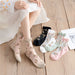 A person wearing sheer floral socks sits on a wooden floor, with four pairs of Lacatang Shop's Vintage Floral Lace Ruffle Socks in Kawaii Harajuku style beside them, in black, white, mint green, and pink. A fashion magazine lies nearby.