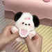 A person holds the Adorable Plush Puppy Keychain by Lacatang Shop, shaped like a small animal with black ears and a pink checkered bow. In the background is a light pink purse with a button clasp on a striped surface, making it an ideal bag charm or gift for any occasion.