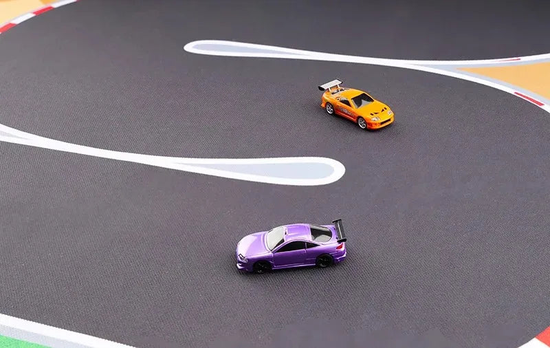 Two 1:76 RC cars, one purple and one orange from Lacatang Shop, speed around the Turbo Racing Rubber Track Mat. The orange car leads on the enhanced grip surface with smooth black curves bordered in white. Options available in 160x90cm or 180x80cm.