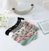 Six pairs of Lacatang Shop's Vintage Floral Lace Ruffle Socks, in colors like black, white, gray, pink, beige, and green, are artfully arranged on a round white table alongside daisies and a book titled "In Spain.