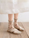 A person wearing a white skirt, Lacatang Shop's Vintage Floral Lace Ruffle Socks, and light beige cross-strap sandals stands on a wooden floor. The socks are Kawaii Harajuku-style with small pink and green floral designs, exuding vintage charm.