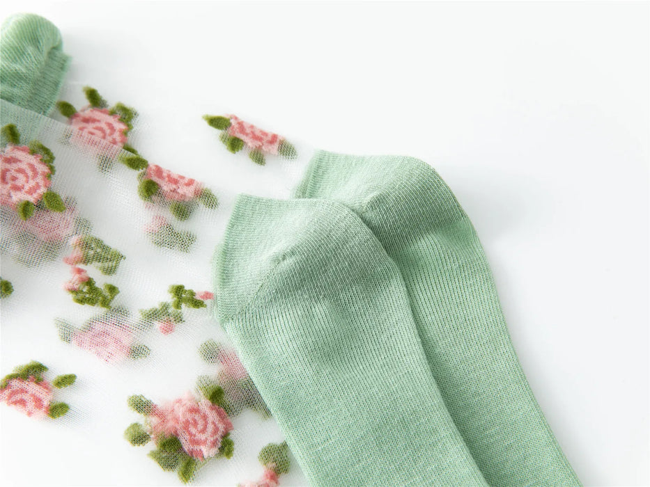 A pair of Vintage Floral Lace Ruffle Socks from Lacatang Shop features a transparent silk design with pink roses, green leaves, and solid light green toes and cuffs, offering a Kawaii Harajuku style. The socks are displayed on a white surface.