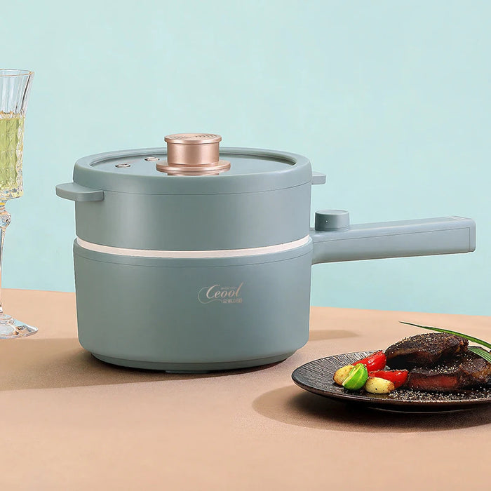 A teal-colored 2024 New Portable 1.5L Multi-function Pot Rice Cooker Electric Cooking Stir Frying Noodle Pot Mini Hot Pot from Lacatang Shop, featuring a long handle and a lid, sits on a beige table next to a transparent, ornate glass filled with a green beverage. Beside the multifunctional electric cooking pot is a black plate with a portion of cooked meat garnished with cherry tomatoes and greens.