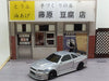 A small silver Lacatang Shop 2.4G RC Drift Car 1/43 4WD Remote Control Car High Speed Four Wheel Drive Radio Controlled Mini Racing Car Model is parked in front of a miniature Japanese tofu shop, adorned with signs above the windows and door. Traditional Japanese text enhances the storefront, and the entire scene is set on a grey and white pavement against a simple background.