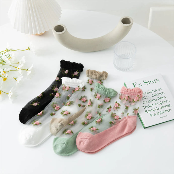 Five pairs of Lacatang Shop's Vintage Floral Lace Ruffle Socks are displayed on a white surface, showcasing Kawaii Harajuku style in black, white, beige, light pink, and green. A vase, glass of water with "EN SPAIN," and white flowers are also visible.