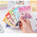 A hand holds several packs of a 100-piece Sanrio Kawaii Sticker Set from AliExpress, featuring Pochacco, Kuromi, Melody, and Hello Kitty. Perfect for student stationery and birthday gifts, these stickers come in various pastel-colored packaging.