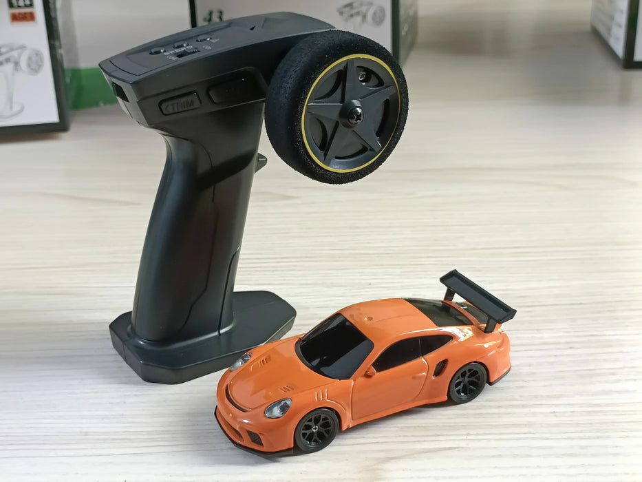 A black handheld RC car controller with a steering wheel and trigger is next to a small, bright orange Lacatang Shop 2.4G RC Drift Car, a high-speed 1/43 scale four-wheel drive toy sports car with a spoiler, on a light wooden surface. The background is blurred with some indistinct objects visible.