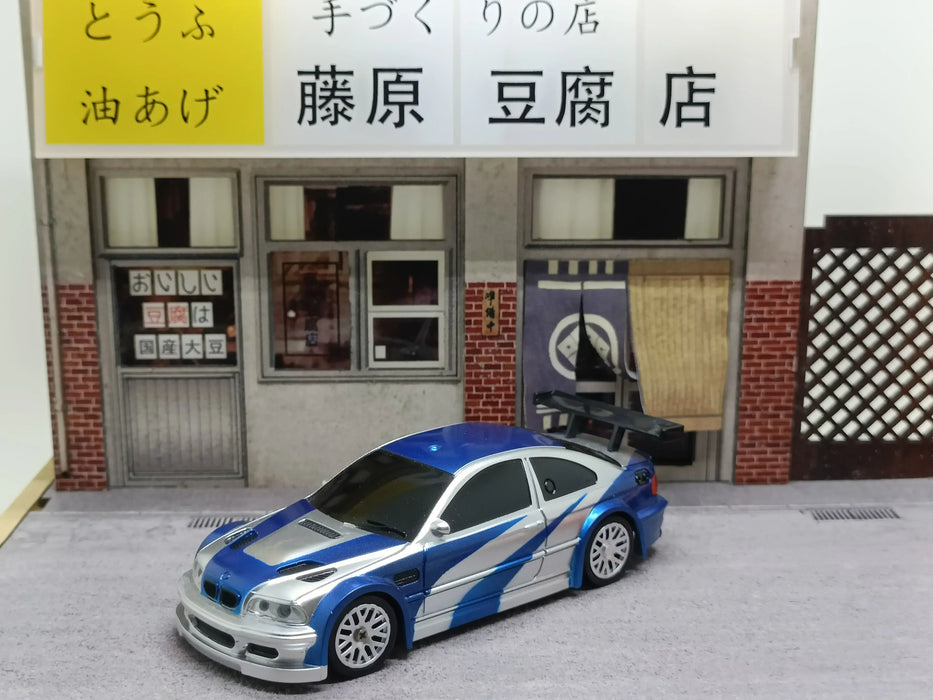 A 2.4G RC Drift Car 1/43 4WD Remote Control Car from Lacatang Shop, featuring a sleek blue and silver design, is parked in front of a carefully crafted miniature Japanese tofu shop. The shop displays signs with Japanese characters and has a quaint fabric curtain draped over the entrance. The model includes detailed storefront windows and a small lattice fence on the right side.