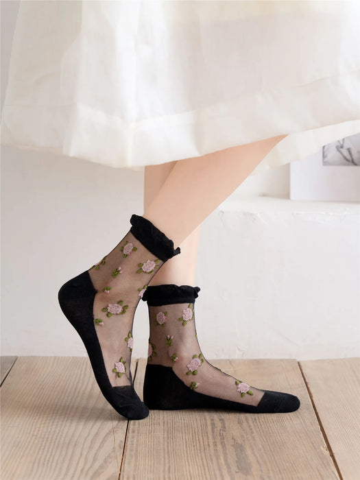 A person in Lacatang Shop's Vintage Floral Lace Ruffle Socks, featuring black cuffs, heels, and toes with pink flower embroidery, stands on a wooden floor. Complemented by a white Kawaii Harajuku-style dress falling to the calves.