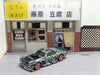 The Lacatang Shop's 2.4G RC Drift Car 1/43, a high-speed, 4WD remote control car featuring a black and white camouflage pattern with brown wheels, is showcased in front of an intricately detailed miniature model of a Japanese tofu shop complete with signs and brickwork. The background includes storefront elements like a door, window, and traditional signage.