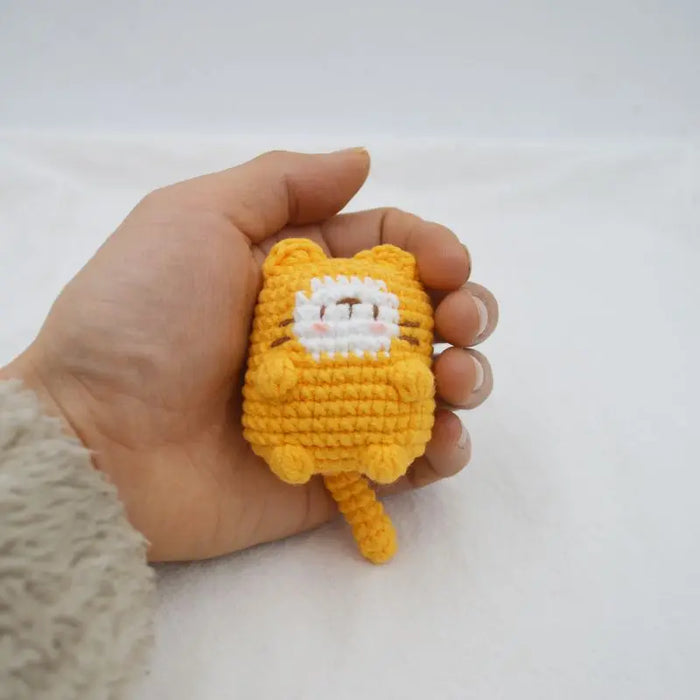 A hand holding a Cute Cat Crochet Keychain from Lacatang Shop, showcasing a small, yellow cat plushie with a white face and brown stripes. The round cat figure features tiny ears, a long tail, and short paws, resembling an adorable knitted keychain. It is set against a plain white background.