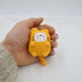 A hand is holding a "Cute Cat Crochet Keychain" from Lacatang Shop, featuring a small, knitted yellow cat with a white face and paws. Resembling an adorable cartoon-style doll, this tiny-tailed and eared keyring comfortably fits in the palm against a plain, white background. The person's fuzzy-sleeved arm is also visible.