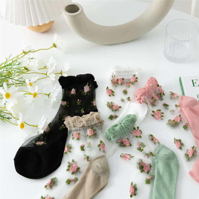 A set of Vintage Floral Lace Ruffle Socks by Lacatang Shop, featuring a Kawaii Harajuku style, is laid out. These ultra-thin transparent socks come in colors like black, white, pink, and green. Nearby are daisies, a modern vase, a glass, and an open book.