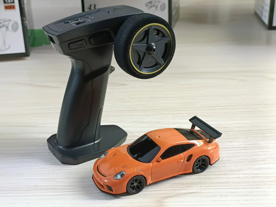 A black 2.4G remote control with a steering wheel and knobs is placed on a light wood surface next to an orange Lacatang Shop 1/43 scale high-speed racing car with a rear spoiler. The background shows the edge of a table and some indistinct items.