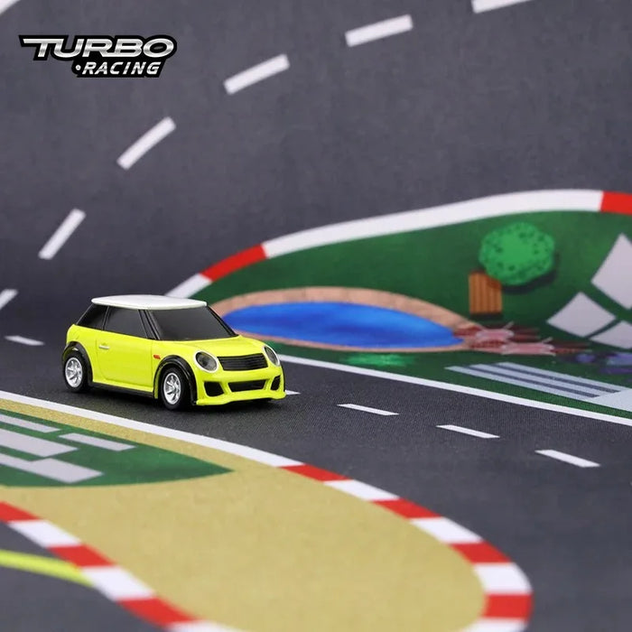 A small yellow toy car sits on Lacatang Shop's Portable Turbo Racing Rubber Mat for 1:76 RC Mini Car Track, featuring a black and white racetrack with winding roads, green patches, and a blue pond, making the car's details pop against this vibrant playground.