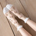 Feet adorned in Lacatang Shop's Vintage Floral Lace Ruffle Socks, featuring sheer pink roses and green leaves, rest on a wooden floor. These Kawaii Harajuku style socks charm with white toes and cuffs.