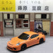 A 2.4G RC Drift Car 1/43 4WD Remote Control Car by Lacatang Shop, sporting a vibrant orange color, is positioned in front of a traditional tofu shop. The sign of the shop displays Japanese characters, and the storefront features a beige and brown color scheme with two large windows and an entrance door. Above the entryway hangs a fabric curtain adorned with a tofu cube design.