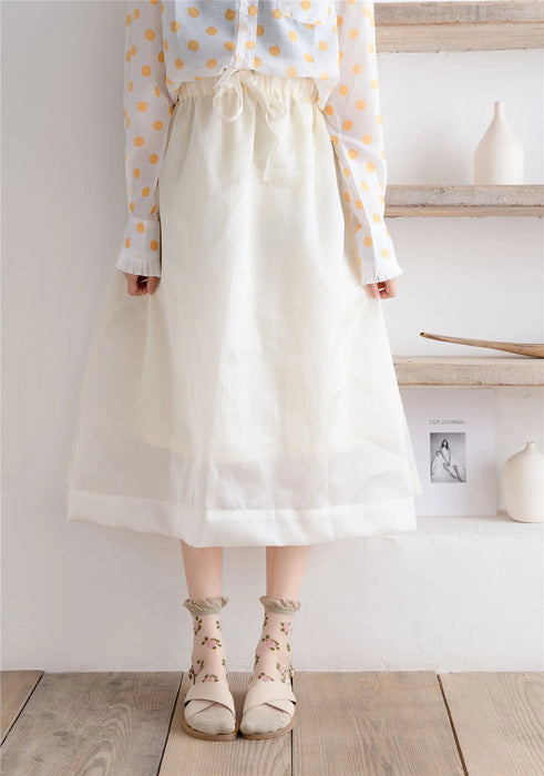 A person in a white polka dot blouse, cream midi skirt, and Lacatang Shop's Vintage Floral Lace Ruffle Socks stands on a wooden floor. Nearby, a shelf holds minimalist decor with a framed photo, book, and vases.