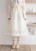 A person in a white polka dot blouse, cream midi skirt, and Lacatang Shop's Vintage Floral Lace Ruffle Socks stands on a wooden floor. Nearby, a shelf holds minimalist decor with a framed photo, book, and vases.