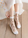 A person in a white dress sits on a wooden floor, adjusting their transparent Kawaii Harajuku style socks from Lacatang Shop. The Vintage Floral Lace Ruffle Socks feature pink floral embroidery and white cuffs. Their dress has sheer sleeves with orange polka dots, adding whimsy to the scene.