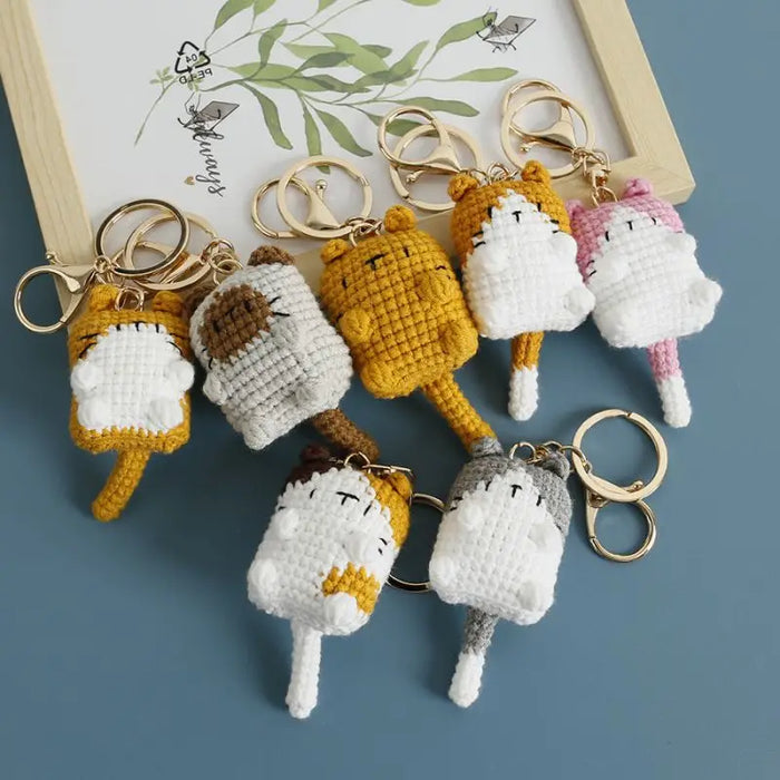 Displayed on a surface, the Cute Cat Crochet Keychains from Lacatang Shop, featuring knitting cartoon cat dolls in various colors like yellow, grey, pink, and white. These kawaii small cat knitted keychains are linked to gold zinc alloy key rings and placed near framed artwork, adding a whimsical touch to your car keys or bag as pendants.