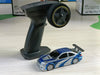 A Lacatang Shop 2.4G RC Drift Car 1/43 4WD Remote Control Car High Speed Four Wheel Drive Radio Controlled Mini Racing Car, featuring a black remote control with a steering wheel and buttons, alongside a small blue racing car with silver detailing, racing stripes, and a spoiler on a light-colored surface.