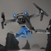 The Yellow Pandora Ninja Dragon Phantom X HD Dual Camera Smart Quadcopter Drone, featuring a sleek grey body with blue lights and foldable arms, soars above a misty, rocky coastal landscape. Its dual cameras capture the scenic view below as dark, jagged rock formations protrude from the water, creating a dramatic backdrop.