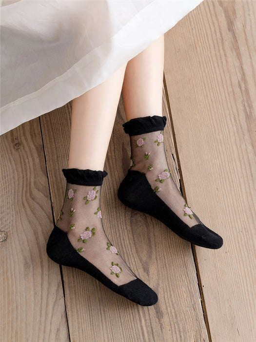 A person sits on a wooden floor displaying Lacatang Shop's Vintage Floral Lace Ruffle Socks, which are Kawaii Harajuku style ultra-thin transparent crystal silk socks with embroidered pink flowers and green leaves. The solid black heels and toes enhance their charm beneath a partially visible white skirt.