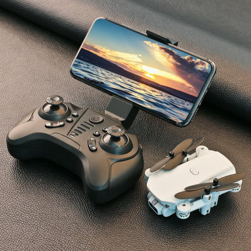 A compact Blue Charlie 4K HD Wide Angle Camera Mini Drone sits next to a remote control with a smartphone mounted on top, displaying a scenic sunset over water. The drone is white with foldable propellers, while the black controller features multiple buttons and joysticks.